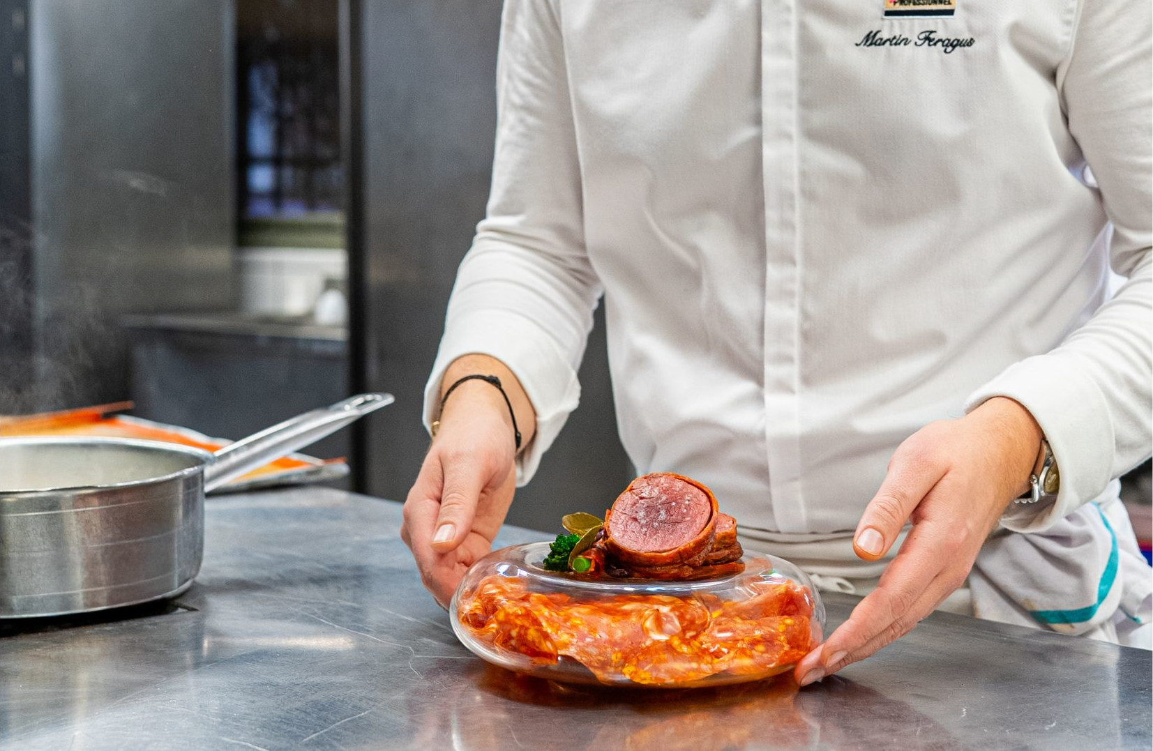 Close-up of a chef presenting a meat dish with red sauce and garnish, with a saucepan on a stainless-steel countertop in a pro kitchen.