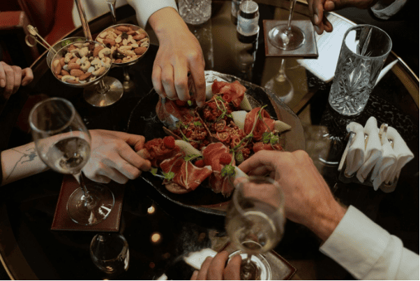 Vue rapprochée d’une table partagée au restaurant avec jambon cru, mains se servant, pain, verres et ambiance conviviale.